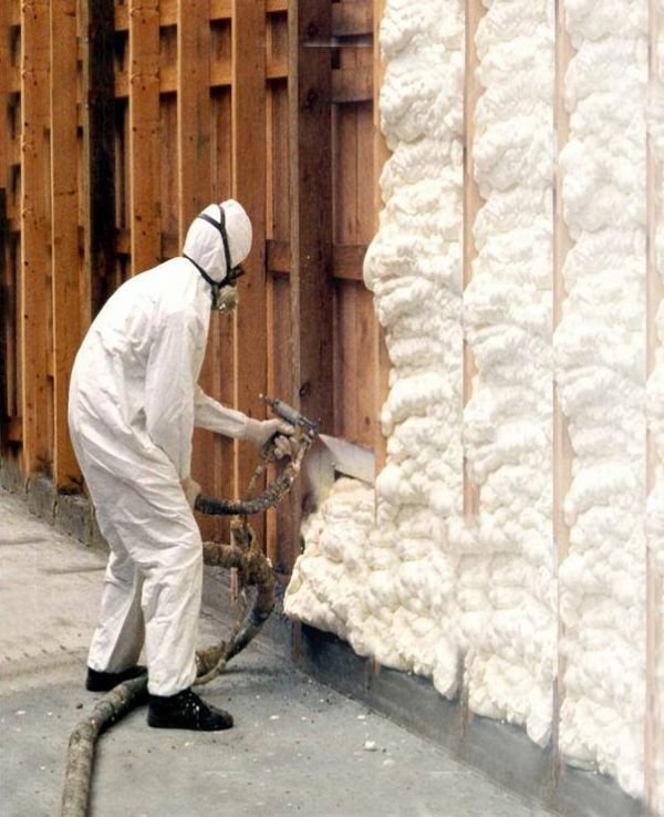 A man in white protective gear applies foam insulation using a spray nozzle in a construction setting.