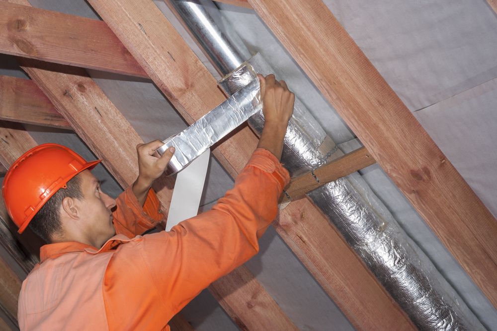 A person wearing an orange shirt and hard hat is applying tape to a silver duct in the attic, surrounded by wooden beams.