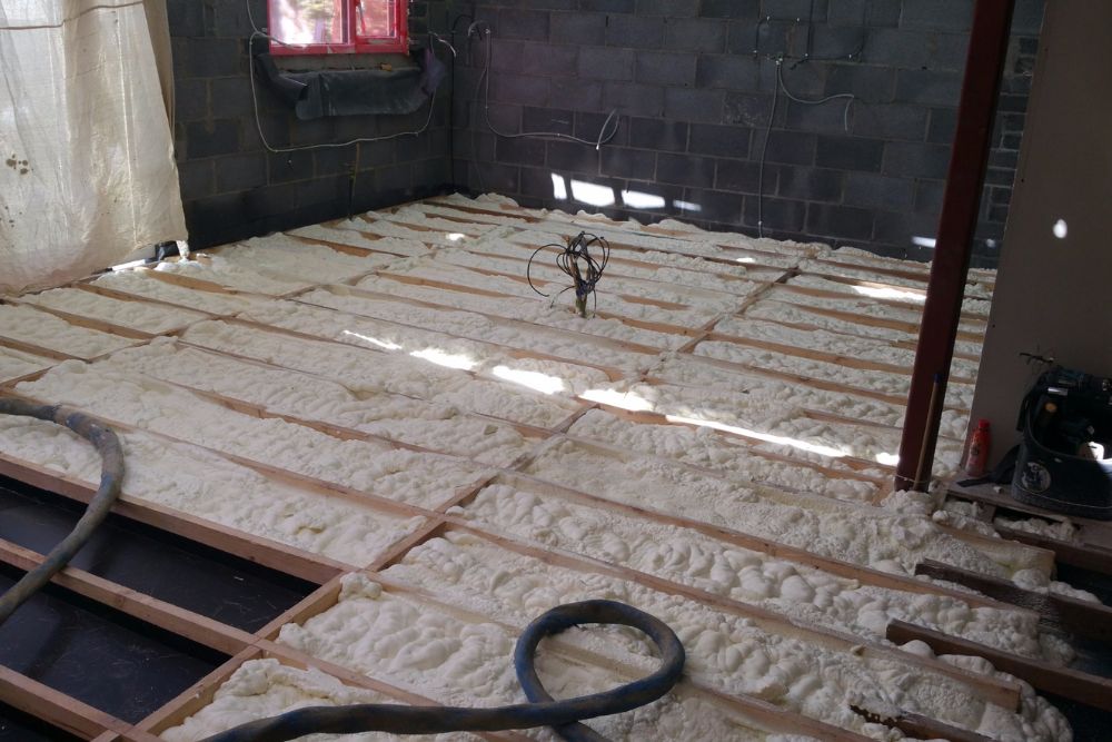 Insulated flooring in a construction area, with foam insulation laid between wooden beams and a vacuum hose on the ground.