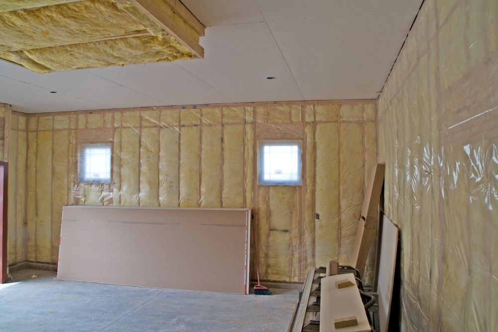 Interior of a partially finished room showing insulation on walls and ceiling, with windows and construction materials on the floor.