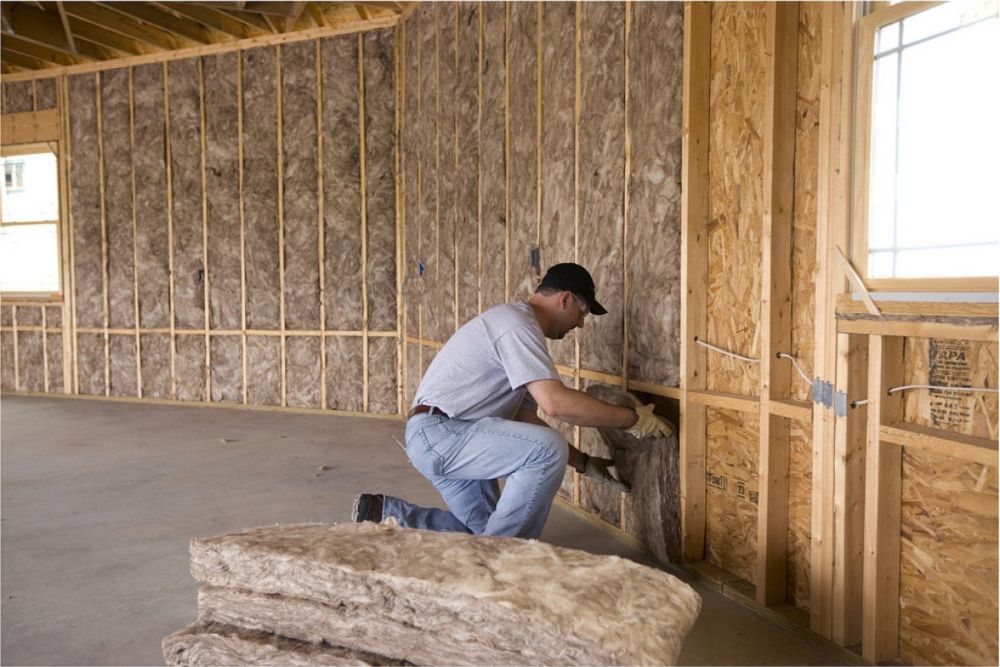 A man works on a wooden wall frame, installing insulation in a partially built house, with insulation batts positioned nearby.