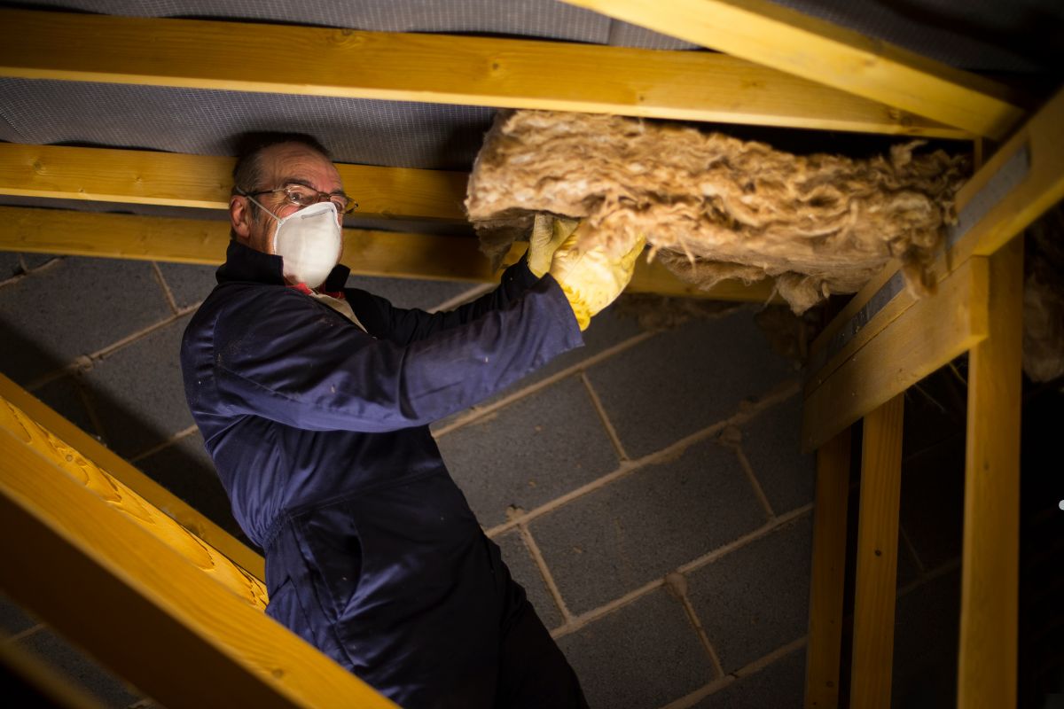  A man in a mask and gloves installs insulation on a roof, ensuring safety while performing his work.