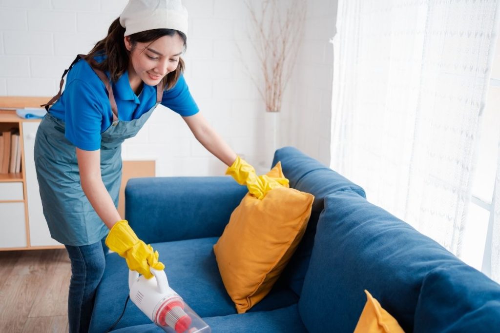 A woman wearing a blue shirt and yellow gloves is cleaning a blue couch with a cloth.